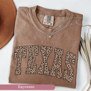 May include: A brown Comfort Colors t-shirt with the word "TEXAS" in large leopard print letters. The shirt has a crew neck and short sleeves. A silver necklace with a small pendant is draped across the shirt.