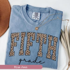 May include: A light blue t-shirt with the word "FIFTH" in large, black outlined letters with a leopard print pattern. The word "grade" is written in cursive below the word "FIFTH". The t-shirt has a tag that reads "COMFORT COLORS".