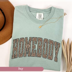 May include: A light green t-shirt with the word "HOMEBODY" in a leopard print design. The shirt has a crew neck and short sleeves. A gold necklace with a pendant is draped on the shirt. The word "Bay" is at the bottom.