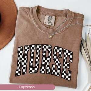 May include: Espresso brown t-shirt with a chequered "NURSE" graphic. The shirt is made of a soft, comfortable fabric and has a relaxed fit.