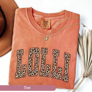 May include: A coral orange t-shirt with the word "LOLLI" printed in large, bold letters. The letters are filled with a leopard print pattern. The shirt has a distressed look and is made of a soft, comfortable fabric.