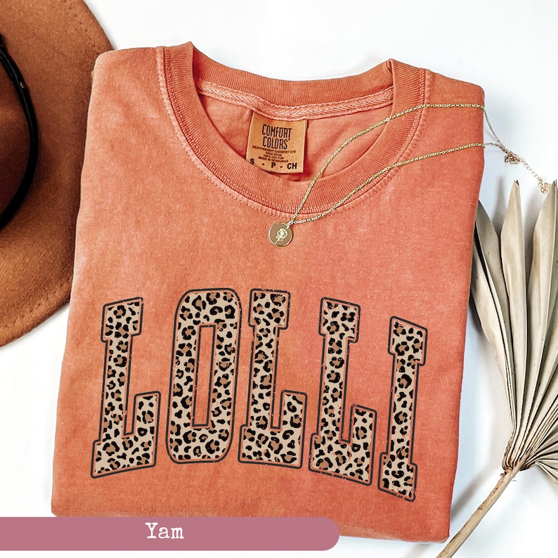 May include: A coral orange t-shirt with the word "LOLLI" printed in large, bold letters. The letters are filled with a leopard print pattern. The shirt has a distressed look and is made of a soft, comfortable fabric.