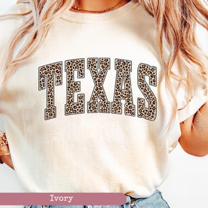 May include: Ivory-colored sweatshirt with the word "TEXAS" in large, block letters. The letters are filled with a leopard print pattern. The sweatshirt has a crew neck and short sleeves.