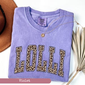 May include: A purple t-shirt with the word "LOLLI" printed in a leopard print design. The shirt is made by Comfort Colors.