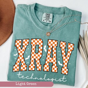 May include: Light green t-shirt with the words "XRAY technologist" in white script. The word "XRAY" is in large orange and white checkered letters, with small bat and star graphics.