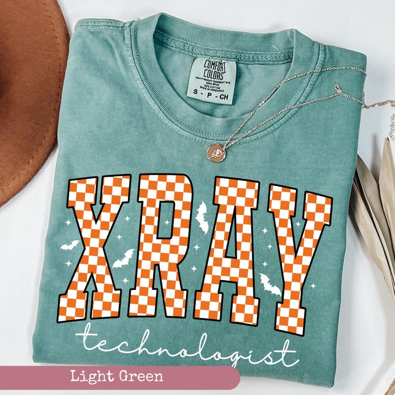 May include: Light green t-shirt with the words "XRAY technologist" in white script. The word "XRAY" is in large orange and white checkered letters, with small bat and star graphics.