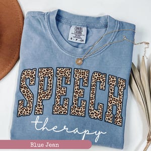 May include: A blue jean colored short-sleeve t-shirt with a crew neckline. The shirt features the word SPEECH in large block letters with a brown and black leopard print pattern, and the word therapy in white cursive script underneath.