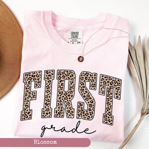 May include: Pink t-shirt with the word "FIRST" in leopard print and the words "grade" in black script below. The shirt has a tag that says "Comfort Colors".