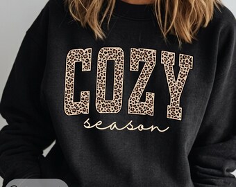 May include: A black crewneck sweatshirt featuring the word COZY in large block letters with a brown and black leopard print pattern. Below the word COZY, the word season is printed in a thin, light-colored cursive script font.