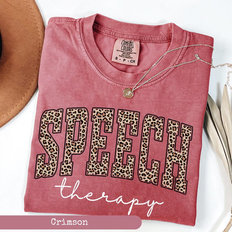 May include: A crimson short-sleeve t-shirt with a crew neckline. The front features the word SPEECH in large block letters with a brown and black leopard print pattern, and the word therapy in white cursive script underneath the block text.