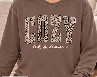 May include: A brown crewneck sweatshirt featuring the word COZY in large, block-style letters with a tan and black leopard print pattern. Below the word COZY, the word season is printed in a thin, white, cursive script font.