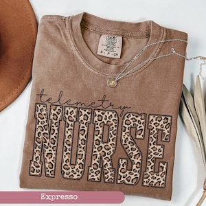 May include: A brown t-shirt with the words "telemetry NURSE" in a leopard print design. The shirt has a crew neck and short sleeves. A silver necklace with a small pendant is draped across the shirt.