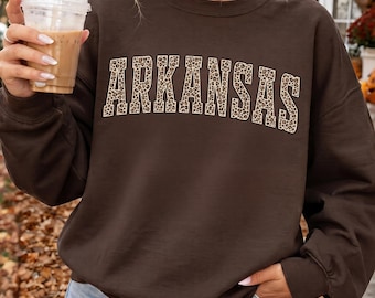 May include: A dark chocolate brown crewneck sweatshirt featuring the word ARKANSAS printed across the chest in a bold, arched collegiate font. The letters have a tan and black leopard print pattern throughout the entire text design.