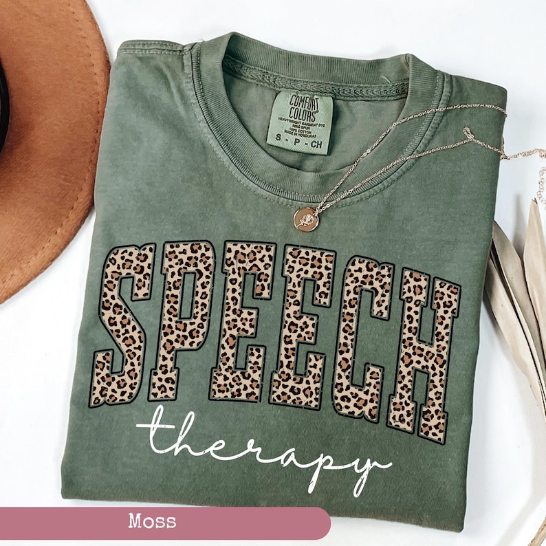 May include: A moss green t-shirt featuring the word SPEECH in large block letters with a brown and black leopard print pattern, followed by the word therapy in white cursive script. The inner neck label reads COMFORT COLORS 100% COTTON.