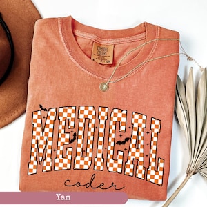 May include: A peach-colored t-shirt with the words "MEDICAL coder" in a checkered orange and white design. The shirt features small bat graphics and a gold necklace. The shirt is from Comfort Colors.