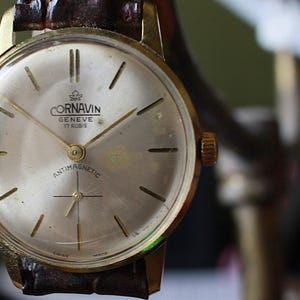 May include: Close-up of a vintage Cornavin Geneve 17 Rubis wristwatch. The watch has a gold-tone case, a light-colored face with gold hands and markers, and a brown leather strap. The face includes the text "ANTIMAGNETIC".