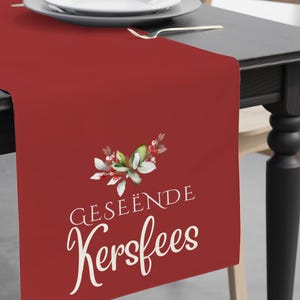 May include: A red table runner with the text "GESEËNDE Kersfees" and a floral design. The runner is on a black table, with a white plate and silverware. A vase with flowers is on the table.