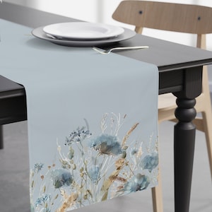 Puede incluir: Un camino de mesa azul claro con un diseño floral de acuarela con flores y hojas azules, marrones y blancas. El camino de mesa está sobre una mesa negra con un plato blanco y un tenedor plateado.