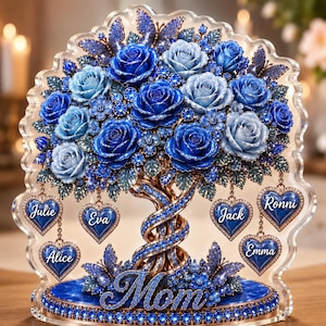 Peut inclure: Un présentoir décoratif en acrylique représentant un arbre avec des roses bleues et des papillons. La base porte le mot Mom en écriture cursive. Six breloques en forme de cœur bleu sont suspendues aux branches avec les prénoms Julie, Eva, Alice, Jack, Ronni et Emma.