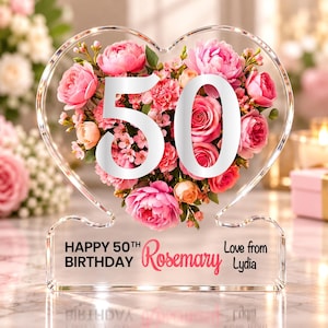 Op de afbeelding: Een hartvormige, doorzichtige acrylplaat met een groot wit getal 50 omringd door roze en perzikkleurige bloemen. De voet bevat de gedrukte tekst: HAPPY 50TH BIRTHDAY Rosemary Love from Lydia in zwarte en roze letters op het oppervlak.