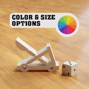Puede incluir: Una catapulta de juguete plateada con un dado blanco a su lado. La imagen incluye el texto "COLOR & SIZE OPTIONS" y una rueda de colores.
