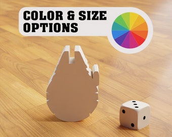 May include: A white, 3D printed model of the Millennium Falcon spaceship from Star Wars. The model is sitting on a wooden surface. A white die with black dots is sitting to the right of the model. A white sign above the model reads "COLOR & SIZE OPTIONS" with a rainbow color wheel to the right of the text.
