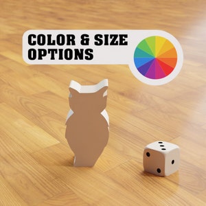 Könnte beinhalten: Eine weiße Eulenfigur und ein weißer Würfel mit schwarzen Punkten auf einer Holzoberfläche. Der Text "COLOR & SIZE OPTIONS" befindet sich über der Eule und ein Farbkreis befindet sich rechts von der Eule.