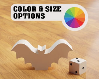 Peut inclure: Une découpe de chauve-souris blanche avec un dé blanc sur un sol en bois. Le texte "COLOR & SIZE OPTIONS" est sur une étiquette blanche avec une roue de couleurs arc-en-ciel.