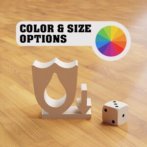 Puede incluir: Un objeto blanco en forma de escudo con un recorte en forma de gota de agua, una flecha blanca apuntando hacia abajo y un dado blanco con puntos negros sobre una superficie de madera. El texto "COLOR & SIZE OPTIONS" es visible sobre los objetos.