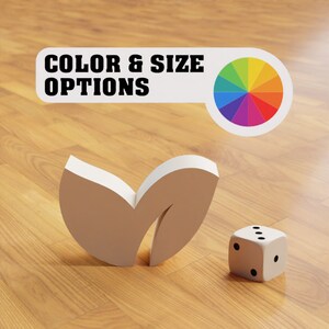 Puede incluir: Un objeto blanco con forma de hoja y un dado blanco con puntos negros sobre una superficie de madera. El texto "COLOR & SIZE OPTIONS" es visible sobre una rueda de colores.