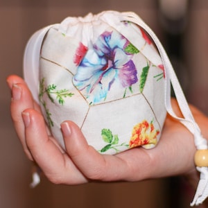 May include: A white drawstring bag with a floral print featuring colorful flowers and gold accents. The bag is held in a hand.