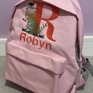 May include: Pink canvas backpack with a zipper closure. The backpack has a front pocket and is embroidered with a zebra, the letter R, and the name Robyn.
