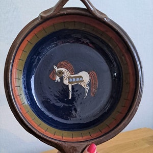 May include: A brown ceramic serving dish with a handle. The dish has a blue center with a stylized white horse painted on it. The rim of the dish is decorated with orange, green, and brown stripes.