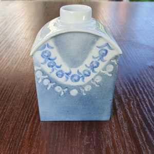 May include: A small, white ceramic vase with a blue and white floral design. The vase is shaped like a house and has a rounded top.