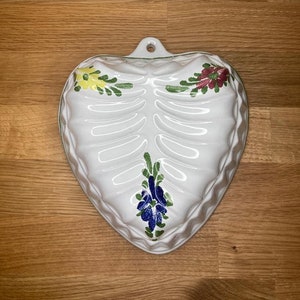 Op de afbeelding: Witte hartvormige keramische vorm met een reliëfdesign en beschilderde bloemdetails in groen, geel, rood en blauw.