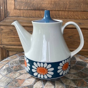 May include: A white ceramic teapot with a blue lid and a band of orange and white daisies on a blue background.