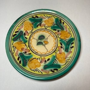 Può includere: Un piccolo piatto rotondo in ceramica con un bordo verde e un centro bianco. Il piatto è decorato con un motivo floreale in tonalità di giallo, verde e marrone.