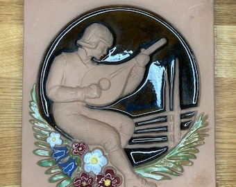 May include: A terracotta tile with a raised relief design of a person playing a lute. The person is wearing a brown hat and a brown tunic. The tile is decorated with green leaves and red, white, and blue flowers.