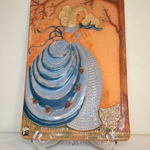 May include: A rectangular ceramic wall plaque depicting a woman in a blue dress holding a blue bird. The artwork has a light orange background with brown tree branches and a brown border. Red roses decorate the dress.
