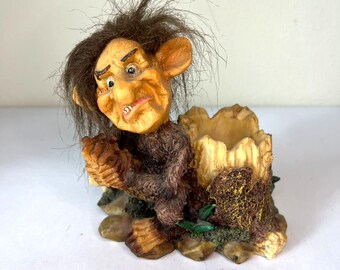 Puede incluir: Una figura caprichosa de un trol con cabello castaño y una expresión gruñona. El trol está ubicado en una maceta de tocón de árbol con una apariencia texturizada y terrosa. La base presenta pequeñas piedras y detalles verdes.