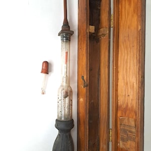 Könnte beinhalten: Ein antikes Hydrometer mit einem Glasrohr, einem Holzgriff und einem bauchigen Sockel, zusammen mit einer separaten Glastropfpipette. Das Hydrometer wird neben einem offenen, gealterten Holzkasten mit Messing Scharnieren und einem Verschluss angezeigt.