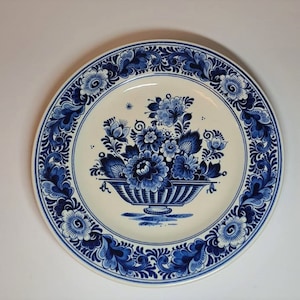 Op de afbeelding: Een wit keramiek bord met een blauw en wit bloemenmotief. Het bord heeft een boeket bloemen in een blauwe en witte vaas, omringd door een decoratieve rand.