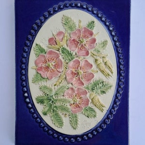 May include: A rectangular ceramic tile with a raised floral design. The central oval features pink flowers with green leaves and yellow buds. The flowers are set against a cream background, framed by a blue border with a beaded edge and a dark blue background.