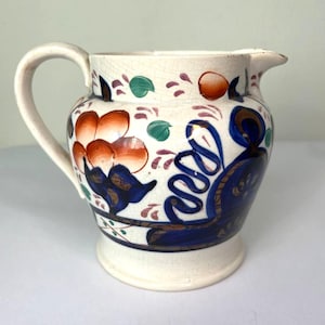 May include: Cream-colored ceramic pitcher with a handle and spout. The pitcher is decorated with orange, blue, green, and brown floral and abstract designs. The base is a solid cream color.