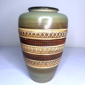Puede incluir: Un jarrón decorativo de cerámica con un cuerpo ancho y un cuello estrecho. El jarrón presenta un degradado de colores, que va desde un borde marrón oscuro hasta un cuerpo verde claro. La sección media tiene bandas horizontales marrones y beige con un diseño de punto de cruz blanco.