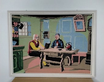 May include: A framed needlepoint picture depicting two people sitting at a table in a room with green walls. The people are wearing colorful clothing and there is a teapot and cups on the table.