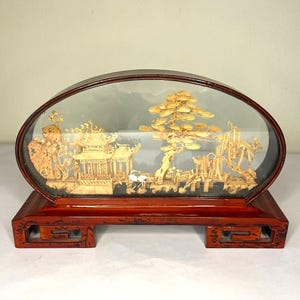 Peut inclure: Un présentoir ovale en bois avec une scène miniature de paysage asiatique. La scène comprend une pagode, des arbres et un pont, tous en matériau brun clair. Le présentoir repose sur une base rectangulaire rouge foncé.