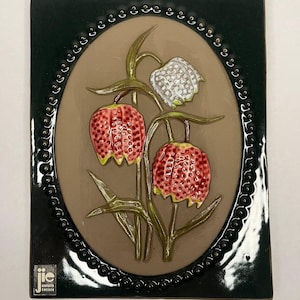 Puede incluir: Un azulejo de cerámica con un diseño en relieve de tres flores rojas y blancas con tallos y hojas verdes. El azulejo está rodeado por un borde negro con un patrón decorativo de pequeños círculos. El azulejo tiene un fondo marrón y el texto "jie carlskrona sweden" está en la esquina inferior izquierda.