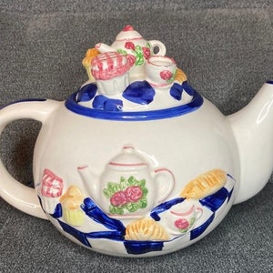 Peut inclure: Théière en céramique blanche avec un motif à carreaux bleus et blancs et un design 3D de cupcakes, tasses à thé et pain. La théière a un couvercle avec un design 3D d'un cupcake et d'une tasse à thé.