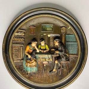 May include: A vintage, circular relief sculpture showing a family scene. The artwork features three figures seated around a table, with detailed clothing and facial expressions. The piece is framed in a dark, ornate border, showcasing a rustic, antique aesthetic.